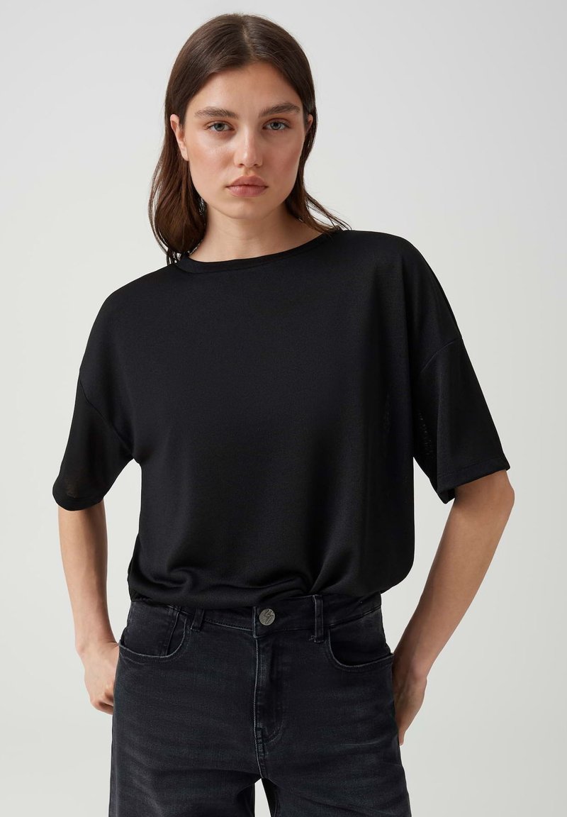 Young woman with straight brown hair wearing a loose black t-shirt and black jeans standing against a plain light background.
