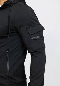 Haut à capuche noir texturé avec une poche zippée sur la poitrine et une poche sur le bras. Doté d'une capuche avec cordon de serrage et d'un design ajusté. Les manches sont lisses.