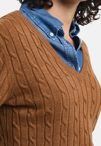 Brauner kabelgestrickter Pullover mit V-Ausschnitt, der über einem blauen Hemd mit Kragen getragen wird. Weiches Stoffdetail sichtbar.