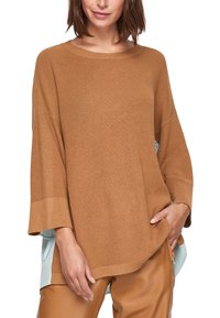 Hnědý pletený svetr s širokými rukávy a volným střihem, překrytý světle modrou košilí. Výrazná textura a kontrastní barevné akcenty.