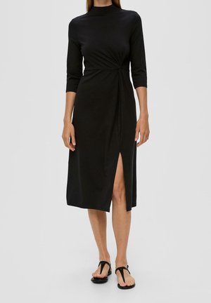 Donna che indossa un vestito midi nero con maniche a tre quarti, spacco laterale e dettaglio nodo, abbinato a sandali neri bassi su sfondo neutro.