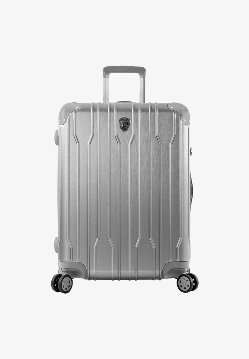 Valise rigide argentée avec des rainures verticales, poignée rétractable et quatre roues noires. Comprend un emblème de logo et des accents argentés.