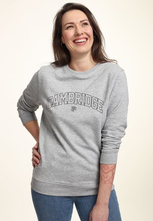 Frau lächelnd, trägt ein graues "Cambridge" Sweatshirt und blaue Jeans, mit einem roten geometrischen Tattoo auf ihrem linken Unterarm.