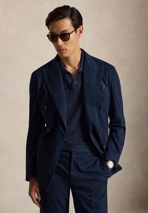 Man draagt een donkerblauwe dubbelgeknoopte blazer met bijpassende broek, een marineblauw gebreid shirt, zonnebril en polshorloge, staand tegen een neutrale achtergrond.