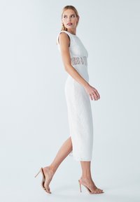 Robe midi en dentelle blanche avec une silhouette ajustée et un panneau en dentelle transparente à la taille. Associée à des sandales à talons hauts couleur nude.