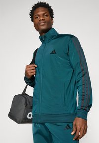 Teal zip-up jack met zwarte zijstrepen en een Adidas-logo. Glad materiaal met een getailleerd ontwerp. Accessoirezak is zwart met witte letters.