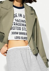 Olivgrön trench coat, vit kortärmad skjorta med svart text och gråa sweatpants med dragsko. Viktiga egenskaper inkluderar knappar och sömmar.