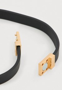 Βραχιόλι με λουράκι από μαύρο δέρμα που διαθέτει ορθογώνιο κούμπωμα σε χρυσή απόχρωση με κυκλικό διακοσμητικό και τέσσερις τρυπητές τρύπες για στερέωση.