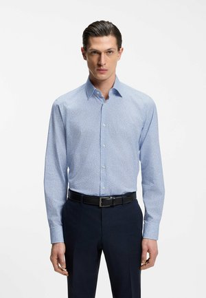 Man draagt een lichtblauw overhemd met patroon, ingestopt in een donkere broek met een zwarte riem, staand tegen een effen witte achtergrond.