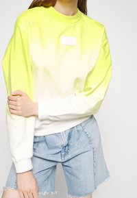 Personne portant un sweat-shirt ombré jaune et blanc avec un short en jean bleu clair, debout devant un fond uni.
