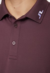 Bordeaux-paita, jossa on teksturoitu kaulus, kaksi nappia ja tyylitelty vaaleansininen logo kauluksessa. Sujuva kangas ja istuva leikkaus.