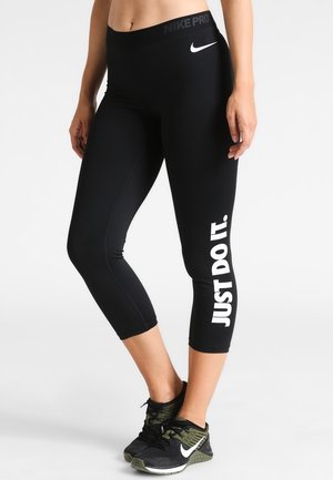 Kobieta nosząca czarne, przycięte legginsy Nike Pro z białym napisem "JUST DO IT." na lewej nodze oraz czarne buty sportowe Nike z białym logo Swoosh.