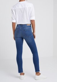 Jeans de mezclilla azul ajustados con un dobladillo deshilachado, bolsillos traseros y una cintura alta, combinados con una camisa blanca de mangas arremangadas y zapatillas blancas.
