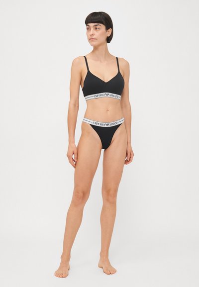 Black bralette and bikini set with a smooth texture. Features logo band in white with "Emporio Armani" text. Adjustable straps, minimal coverage design.