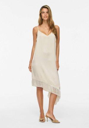Femme aux longs cheveux portant une robe beige asymétrique sans manches avec un ourlet à franges et des sandales beiges à talons, debout sur un fond blanc.