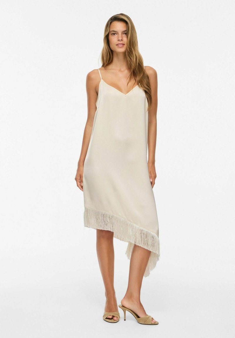 Femme aux longs cheveux portant une robe beige asymétrique sans manches avec un ourlet à franges et des sandales beiges à talons, debout sur un fond blanc.