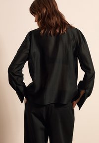 Schwarze durchsichtige Bluse mit langen Ärmeln und Schlitzen an den Manschetten, getragen über schwarzen Hosen. Glatte Textur, lockere Passform und gerader Saum.