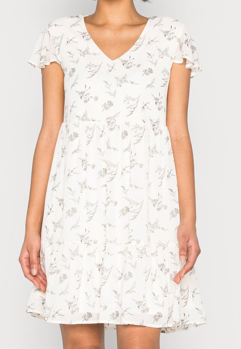 Femme portant une robe blanche à manches courtes avec de petits motifs floraux gris et un col en V, debout devant un fond uni.