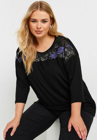 Schwarzes Oberteil mit drei Viertelärmeln, das über ein transparentes, floral besticktes Joch in Lila und Weiß verfügt. Weicher Stoff mit einem lockeren Schnitt.
