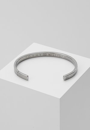 Bracelet manchette ouvert ton argent avec gravure "Tommy Hilfiger" et logo "H" embossé à une extrémité, présenté sur un cube blanc.