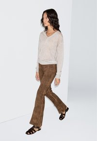 Woman walking wearing a light beige long-sleeve top, brown suede flared pants, and black open-toe sandals against a white background.