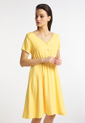 Frau in einem kurzärmligen gelben Kleid mit Knöpfen vorne, stehend und zur Seite schauend vor einem einfarbigen Hintergrund.