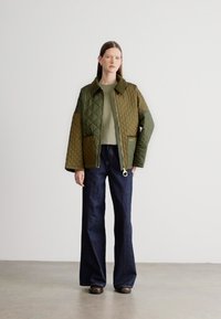 Πράσινο μπουφάν με καπιτονέ σχέδιο, κλείσιμο με φερμουάρ, μεγάλες τσέπες και λεπτομέρεια με κρίκο στη φερμουάρ, συνδυασμένο με σκουρό μπλε καμπάνα τζιν και ένα ανοιχτόχρωμο πουκάμισο.