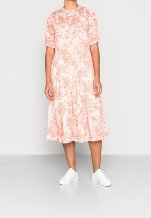 Osoba w krótkiej, sięgającej kolan białej sukience w pomarańczowy wzór kwiatowy oraz białych trampkach, stojąca na gładkiej podłodze.