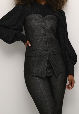 Femme portant un tailleur-pantalon noir texturé avec un blazer boutonné à encolure en cœur et un chemisier transparent à manches longues bouffantes en dessous.