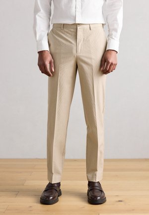 Béžové prispôsobené nohavice s jemnou textúrou, rovný predný diel a bočné vrecká, kombinované s hnedými mokasínmi na svetlej drevenej podlahe.