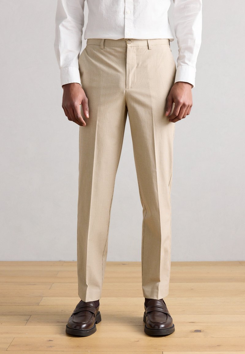 Μπεζ παντελόνι σε ίσια γραμμή με διακριτή υφή, επίπεδη μπροστινή επιφάνεια και πλαϊνές τσέπες, σε συνδυασμό με καφέ λουστρίνι παπούτσια πάνω σε ανοιχτόχρωμο ξύλινο πάτωμα.