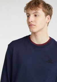 Felpa blu navy in cotone, con scollo a girocollo, maniche lunghe e un piccolo logo nero sul petto. Strato rosso visibile sotto.