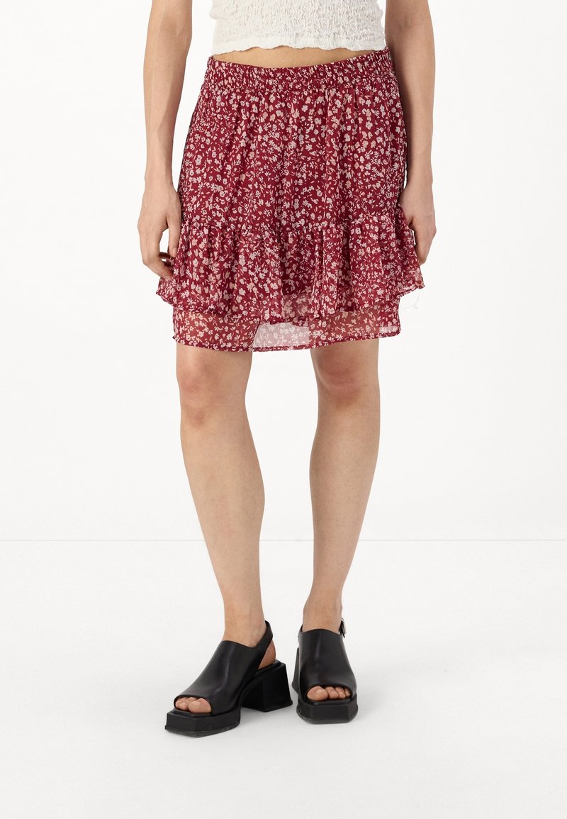 Femme portant une mini-jupe rouge à fleurs avec un ourlet à volants et des sandales à talon large noires à bouts ouverts, debout sur fond blanc.
