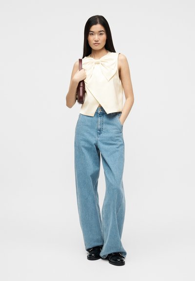 Νεαρή γυναίκα που φοράει μια κρεμ αμάνικη μπλούζα με μεγάλο φιόγκο, ανοιχτόχρωμα γαλάζια παντελόνια με φαρδιά μπατζάκια, μαύρα παπούτσια και κρατάει καφέ τσάντα ώμου.