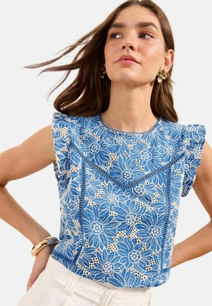 Kobieta ubrana w niebieską kwiatową koronkową bluzkę bez rękawów z falbankami na ramionach oraz białe spodnie, dopełniona złotymi kolczykami w kształcie kwiatów i bransoletką.