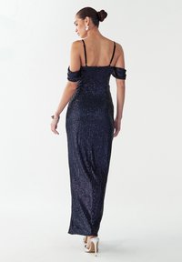 Dolga, oprijeta večerna obleka z naramnicami, ki so spuščenih s ramen, iz mornarsko modrega bleščečega tkanine in elegantne silhuete. Doplačana s čevlji z visokimi petami.