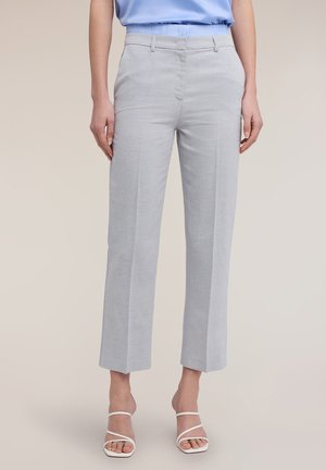 Frau trägt maßgeschneiderte, hellgraue, knöchellange Hose, kombiniert mit weißen, geschnürten Sandaletten mit Absatz und einem hellblauen, in die Hose gesteckten Top.