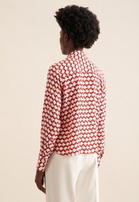 Camicia a maniche lunghe bianca, con un motivo geometrico rosso e nero. Collo tailored, polsini con bottone e vestibilità rilassata.