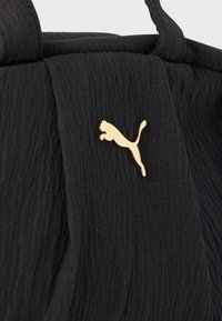 Sac fourre-tout en tissu noir texturé avec une petite épingle logo Puma dorée fixée près du centre supérieur.