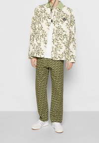 Veste à motif floral blanc avec col vert, associée à un pantalon olive à motif et des baskets blanches. Le tissu semble lisse et léger.