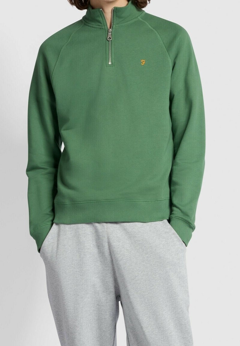 Grüner Pullover mit halbem Reißverschluss am Kragen, Raglanärmeln und einem dezenten goldenen Logo. Der Stoff wirkt glatt, mit einer lockeren Passform und einem gerippten Saum.