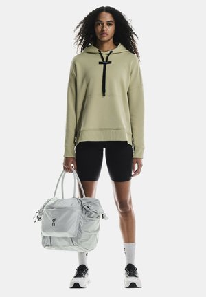 Jasnooliwkowy hoodie z przednią kieszenią, czarnym sznurkiem i ściągaczem, w połączeniu z czarnymi szortami rowerowymi i szarym torbą sportową.