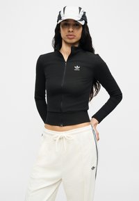 Γυναίκα που φοράει μαύρο μπουφάν Adidas με φερμουάρ, λευκές φόρμες Adidas και μαύρο-άσπρο καπέλο, μπροστά σε απλό ανοιχτόφωτο φόντο.