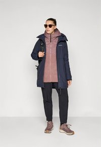 Marineblauwe waterdichte jas met capuchon, roze fleece met rits eronder, zwarte broek en bruine wandelschoenen. Inclusief zonnebril en rugzak.