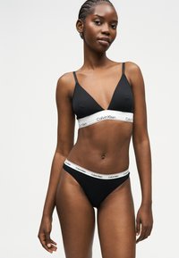 Femme noire portant un bralette Calvin Klein noir et des sous-vêtements assortis avec des bandes élastiques blanches marquées, debout devant un arrière-plan clair uni.