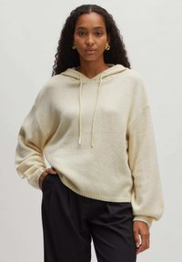 Cremfarbener, übergroßer Hoodie mit Kordelzug, geripptem Saum und strukturiertem Stoff. Kombiniert mit dunklen Hosen.