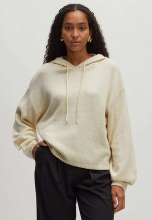 Cremfarbener, übergroßer Hoodie mit Kordelzug, geripptem Saum und strukturiertem Stoff. Kombiniert mit dunklen Hosen.