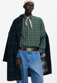 Zielono-niebieska koszula w kratę z guzikami, jasne dżinsy, brązowy pasek ze srebrną klamrą, ciemny płaszcz z wzorem w kratę i jasny beanie.