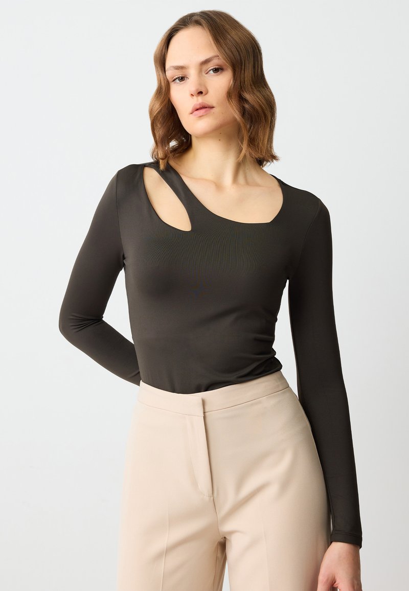 Top lungo colore olivo scuro con un taglio unico su una spalla, tessuto morbido e una silhouette aderente, abbinato a pantaloni beige a vita alta.