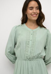 Mint green dress with long sleeves, featuring a button front, lace detailing, and elastic waist for a relaxed fit. Smooth fabric texture.